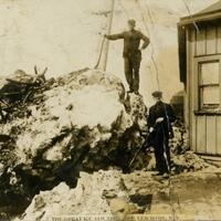 Two men stand on giant piles of ice from an ice jam. A small wooden building is on the right side of the image.