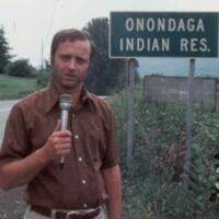 Reporter with microphone in front of sign reading "Onondaga Indian Res."