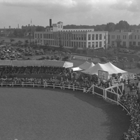Looking southwest from Exposition Park toward Dewey Avenue and the buildings of the City of Rochester's Department of Public Works. In the foreground, crowds surround the horse show arena. Behind the grandstand, cars fill the parking area. The larger white tent in the center has a sign reading "Tea Tent".