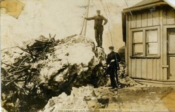 Two men stand on giant piles of ice from an ice jam. A small wooden building is on the right side of the image.